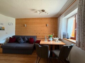 Een woonkamer met een houten wand, een grijze bank met rode kussens, een houten eettafel met stoelen en een raam met gordijnen.