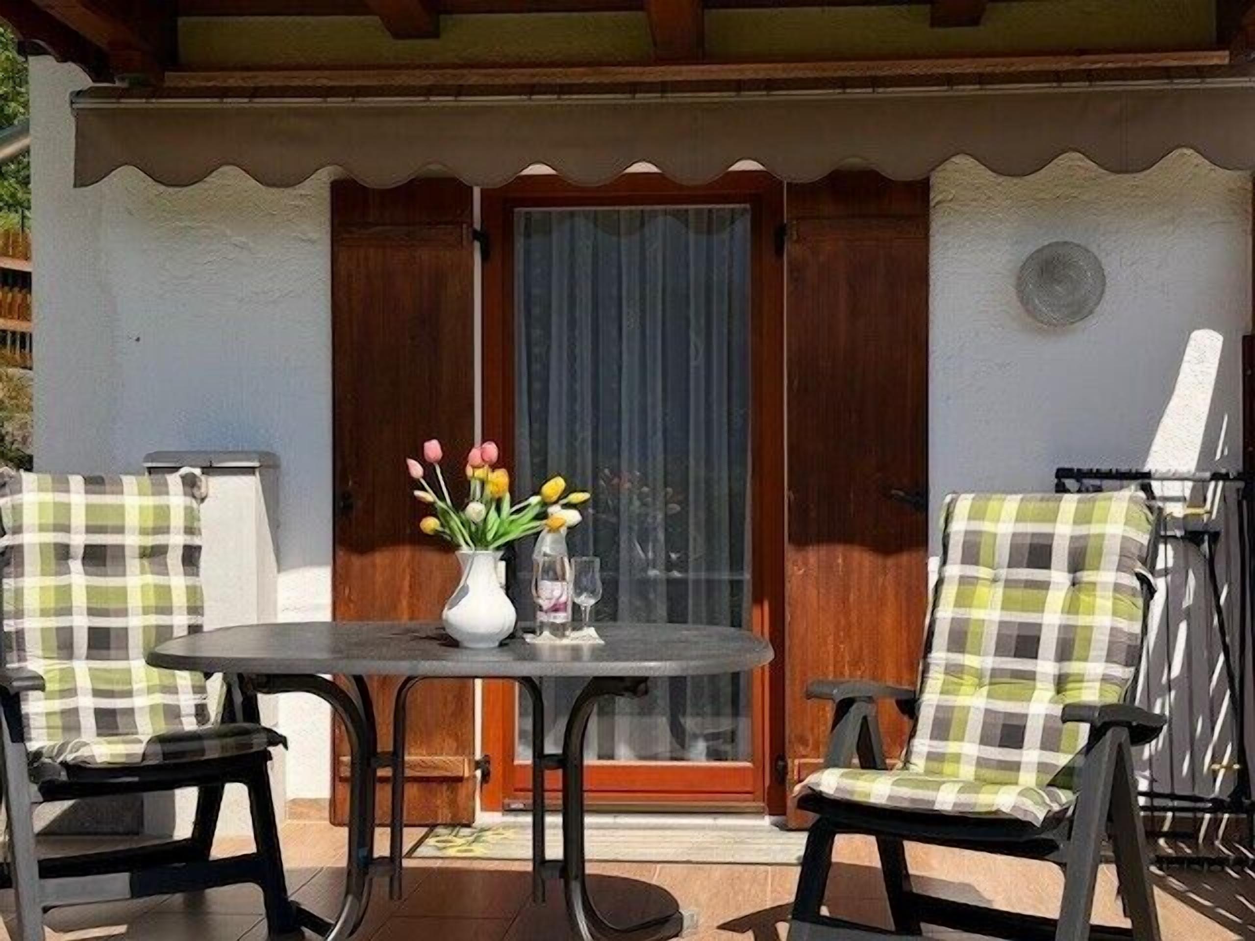 An outdoor seating area with a table and chairs, a vase with flowers, and a wooden door.