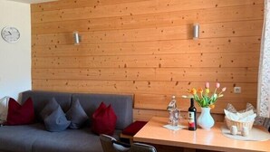 A room with a wooden wall, a grey sofa with red cushions, a table with a bottle, a vase with flowers, and a clock on the wall. - Vacation apartment Zuhaus- 2 persons, 45sqm, terrace with mountain view (Bad Reichenhall)
