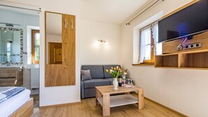 A modern living room with a wooden floor, a grey sofa, a wooden coffee table with a vase of flowers, and a TV mounted on the wall. - Vacation apartment in the farmhouse- 2 persons, 30sqm, balcony with mountain view (Bad Reichenhall)