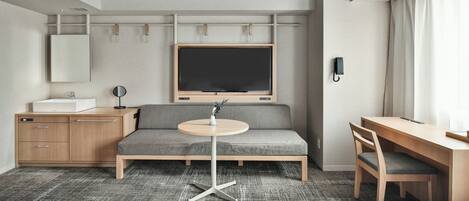 A modern hotel room with a sofa, a desk, a round table, a TV, a mirror, and a phone.