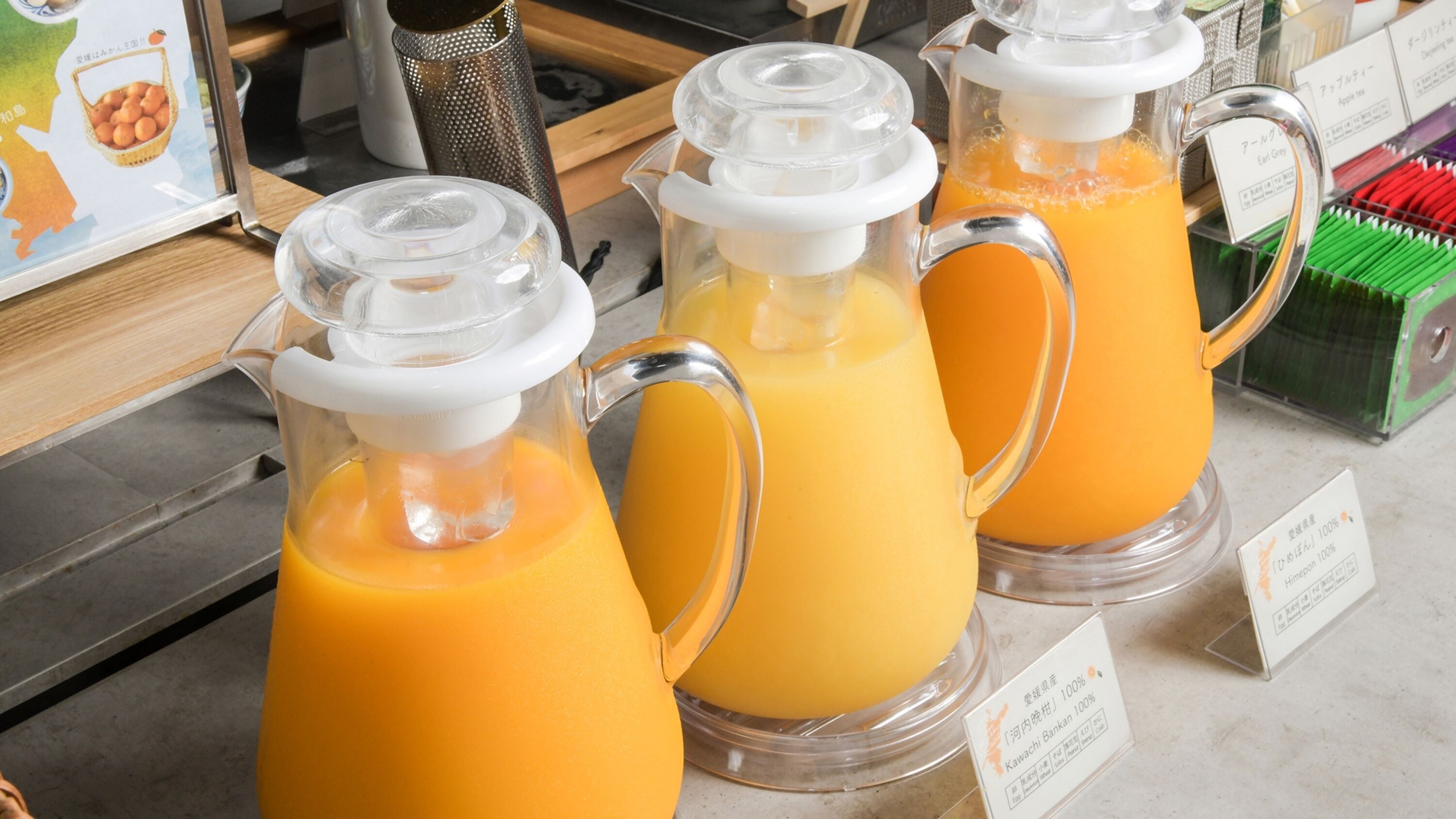 Three glass dispensers of orange juice with white lids and handles, placed on a counter with labels indicating the juice type.