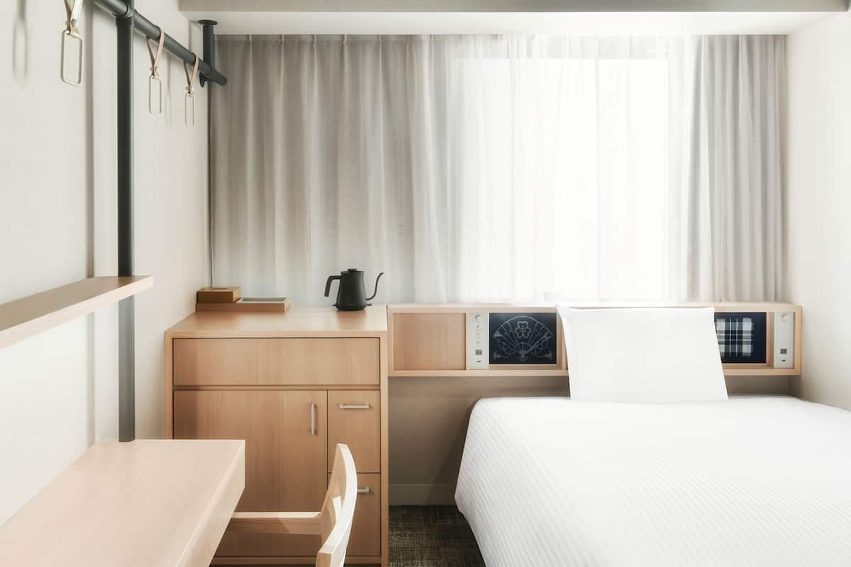 A hotel room with a bed, a desk, a chair, a small table, a kettle, and a shelf with a picture frame and a clock.