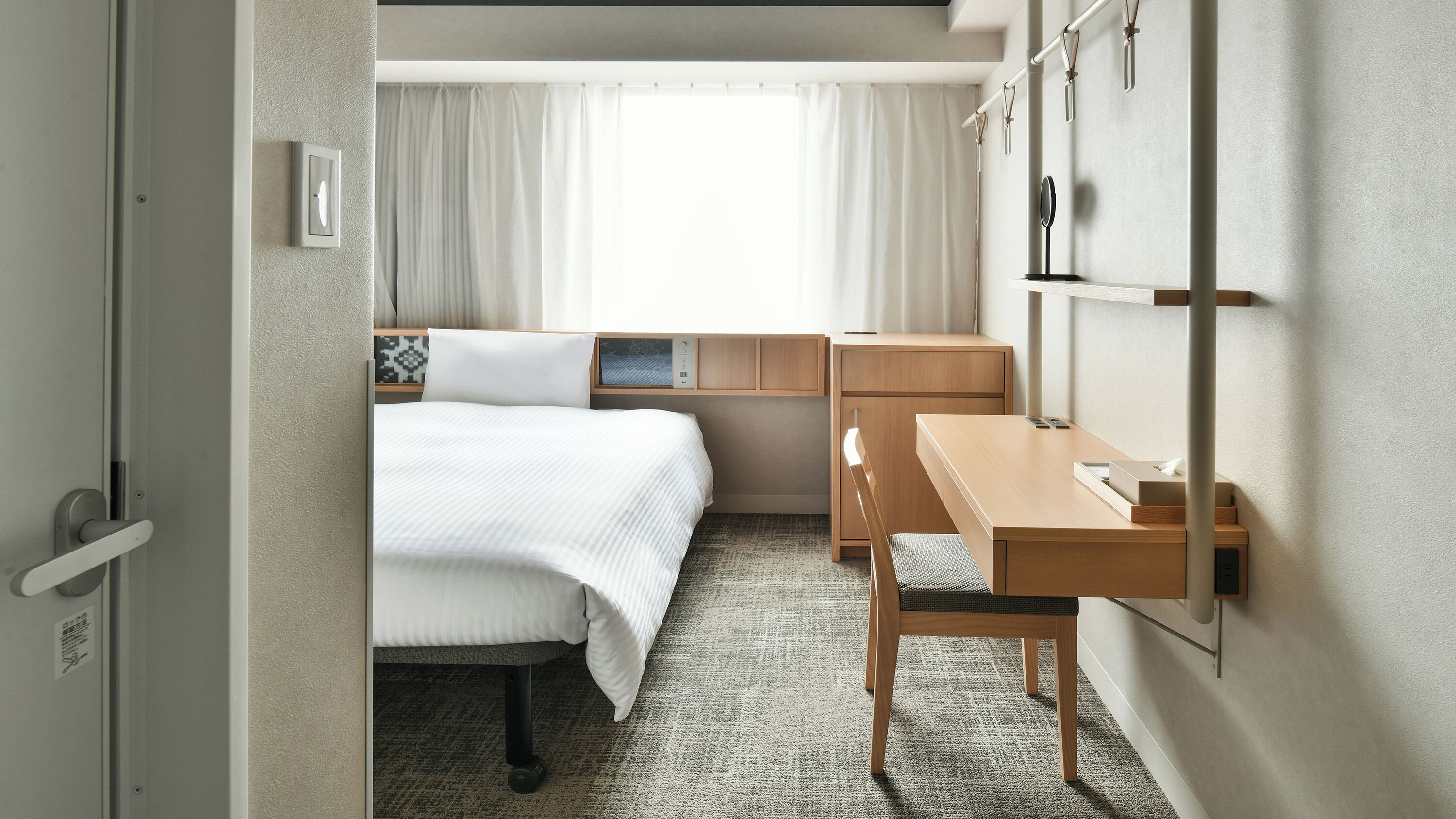 A hotel room with a bed, a desk, a chair, a wardrobe, and a window with curtains.