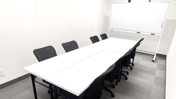 Un salón de conferencias con una larga mesa blanca, seis sillas negras y una pizarra en la pared.