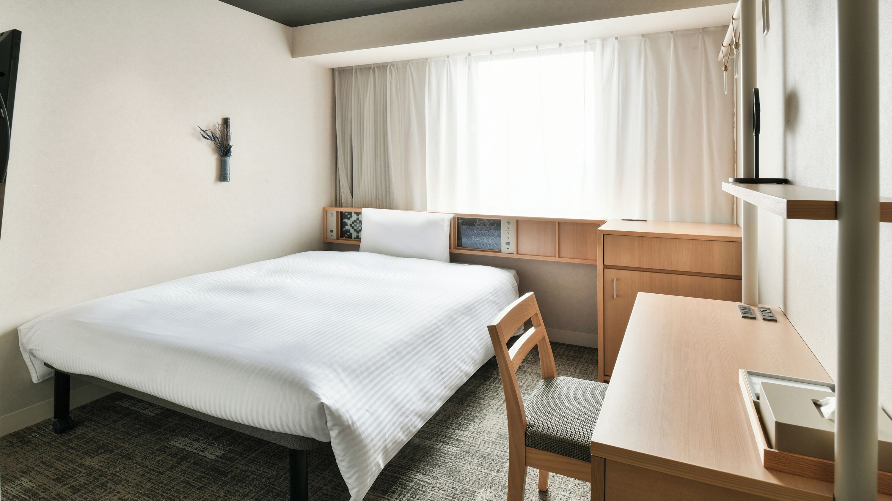 A hotel room with a bed, a desk, a chair, a small table, a window with curtains, and a wall-mounted TV.