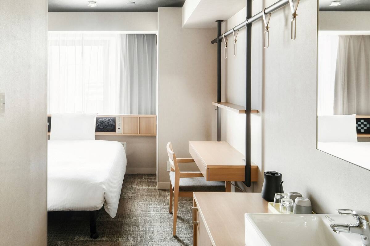 A hotel room with a bed, a desk, a chair, a large mirror, a sink, and a window with curtains.