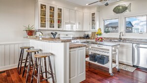 A kitchen with white cabinets, a central island with stools, a ceiling fan, and a view of a house through a window. - "Sandpiper Crossing" - Luxury Oceanfront Home, 4 Bedrooms / 3.5 Bathrooms (Holden Beach)