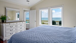 A bedroom with a large bed, a dresser, and a view of the ocean through a door. - "Sandpiper Crossing" - Luxury Oceanfront Home, 4 Bedrooms / 3.5 Bathrooms (Holden Beach)