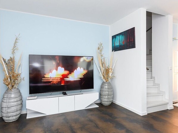 A modern living room with a flat-screen TV on a white entertainment unit, two decorative vases, and a staircase leading to an upper floor. - Vacation cottage Nordseeperle (Dornum)
