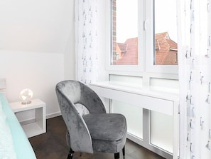 A room with a grey chair by a window, a white bedside table with a globe, and a bed with a light blue cover.