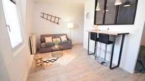 Ein modernes Wohnzimmer mit grauem Sofa, einem kleinen Holzschemel, einem Couchtisch mit Pflanze, einer Stehlampe und einer Bar mit Sitzgelegenheiten.