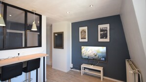 Ein modernes Wohnzimmer mit einer Holztrenne, zwei schwarzen Stühlen, einer weißen Bank, einem an der Wand montierten Fernseher und zwei gerahmten Bildern.