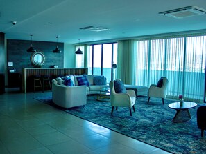Een moderne woonkamer met een bank, fauteuils en een salontafel. Er is een groot raam met gordijnen, een stenen muur en een hangende lamp.