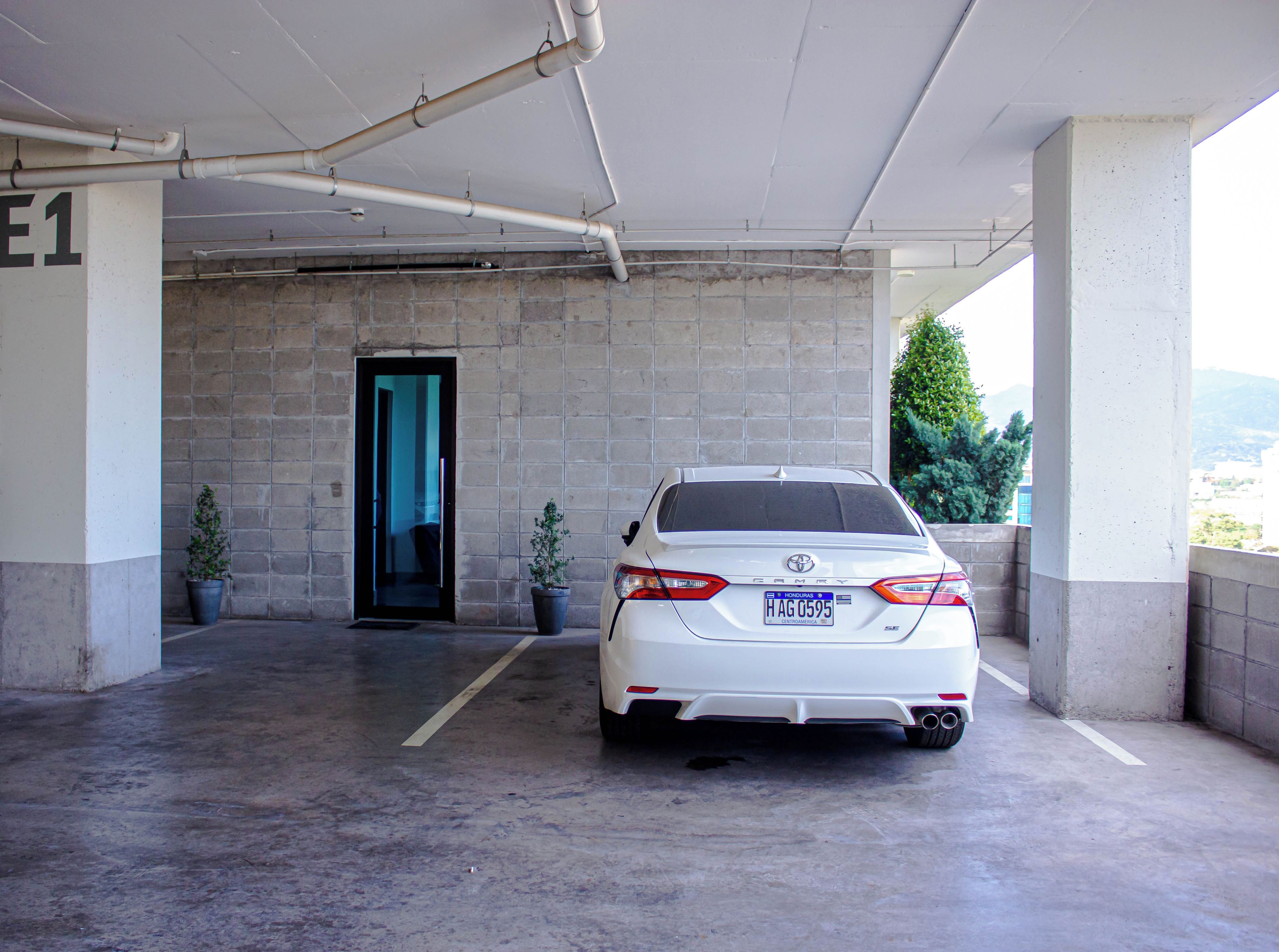 A white car parked in a covered parking area with a concrete floor, a visible door leading to another section, and potted plants.