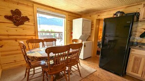 Interior de una cabaña de madera con una mesa de comedor, sillas, un refrigerador negro, una lavadora y una ventana con vistas a la montaña.