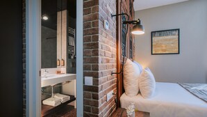 A hotel room with a brick wall, a bed with white bedding, a bedside table with a lamp, and a painting on the wall. - Tbilisee Hotel (Tbilisi)