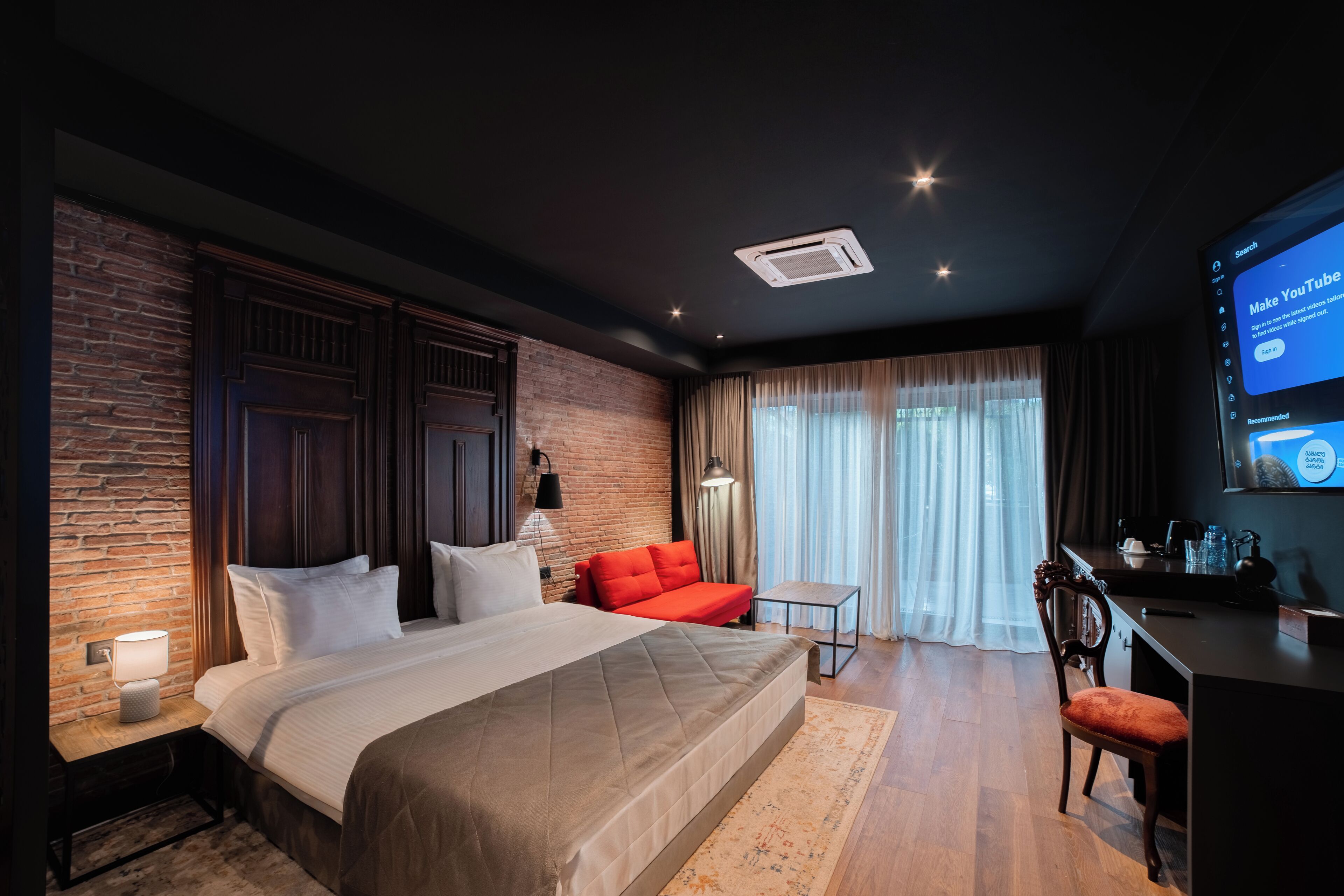 Habitación de hotel moderna con pared de ladrillo decorativa, una cama grande, un sofá rojo, un escritorio y un televisor mostrando la interfaz de YouTube.