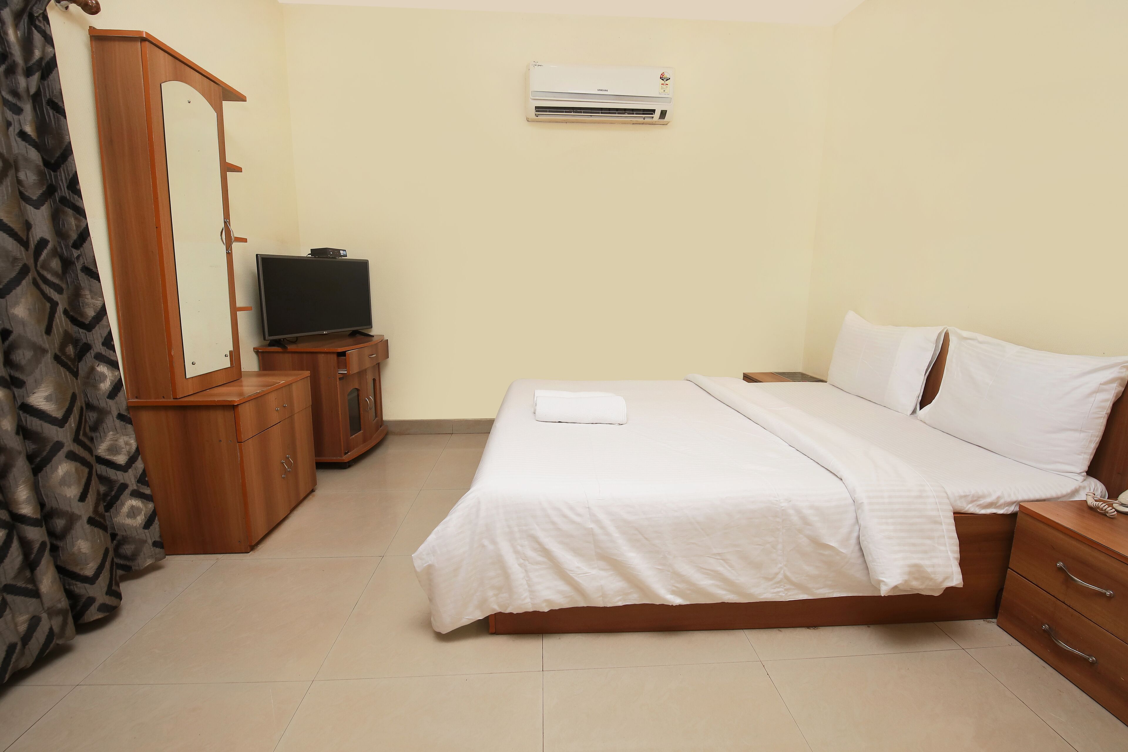 A bedroom with a bed, a TV on a wooden cabinet, a wardrobe, and an air conditioner on the wall.