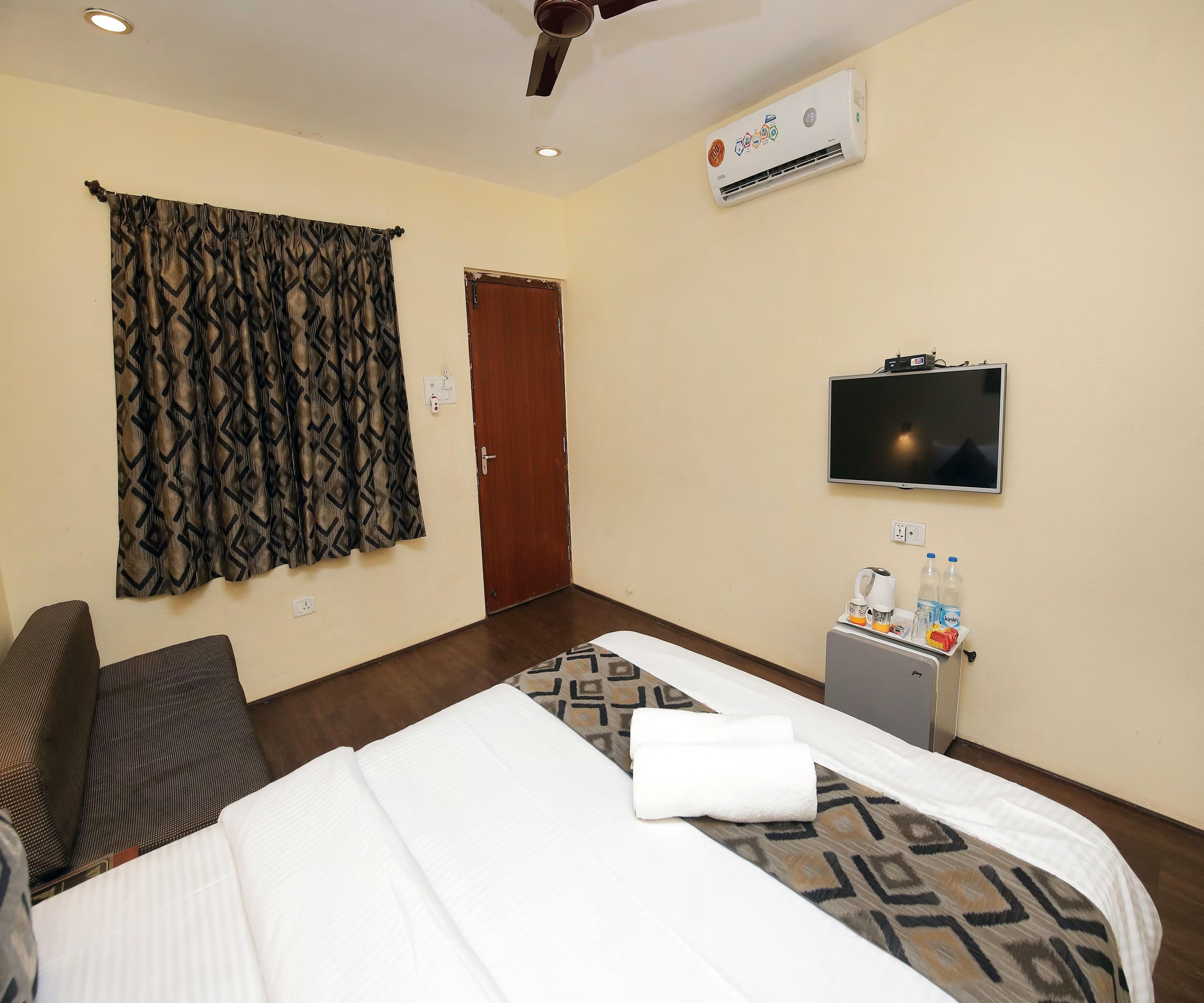 A hotel room with a bed, a TV, a small kitchenette, and a window with curtains.