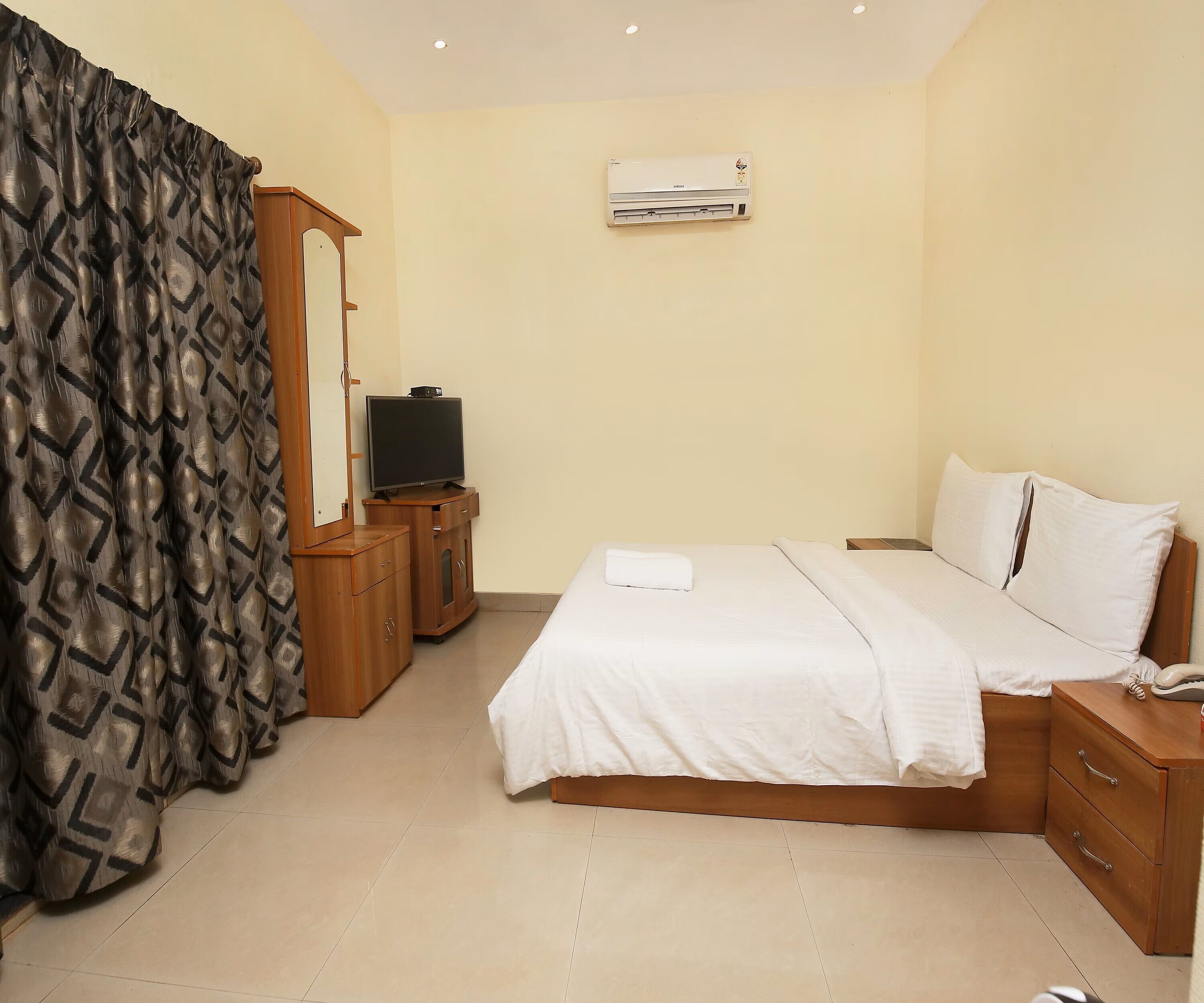 A hotel room with a bed, a television, a wardrobe, a mirror, and an air conditioning unit.