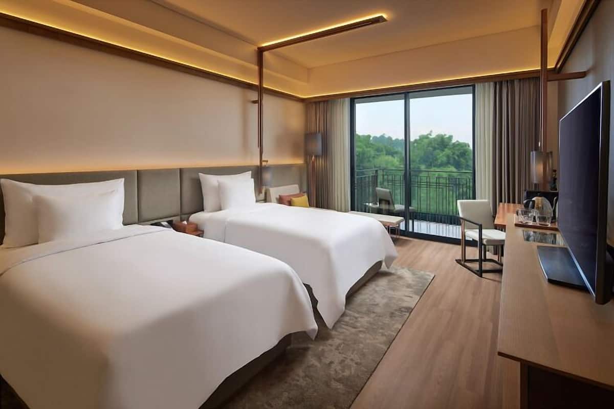 A hotel room with two beds, a desk with a TV, and a balcony with a view of greenery.