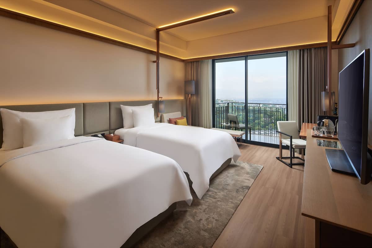 A hotel room with two beds, a desk with a TV, and a balcony with a view.