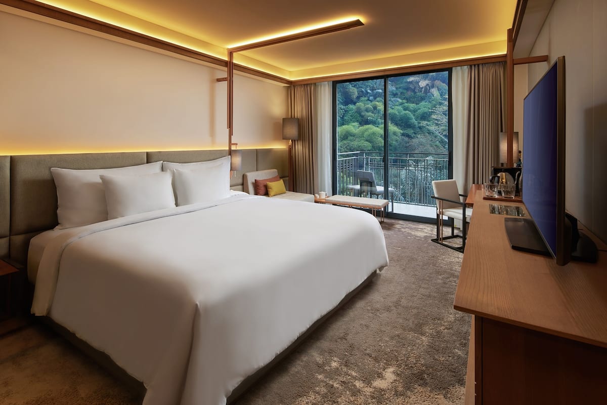 A hotel room with a large bed, a desk with a laptop, a TV, and a balcony with a view of greenery.