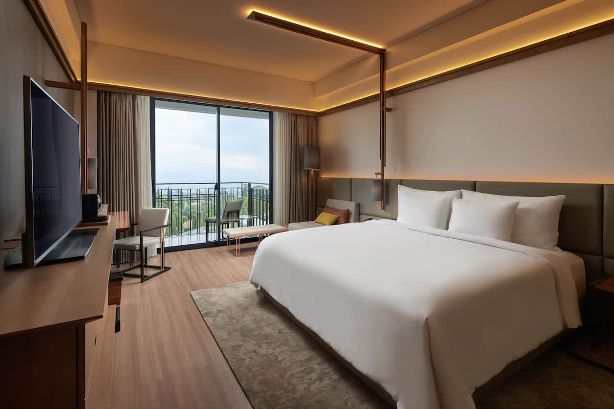 A modern hotel room with a large bed, a desk with a flat-screen TV, a chair, and a balcony with a view.
