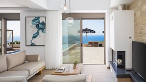 Un salon moderne avec un canapé, une table basse et un téléviseur à écran plat. Un grand tableau orne le mur, et on peut admirer l’océan à travers les portes vitrées.