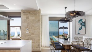 Une cuisine moderne dotée d’un grand îlot central, d’un coin repas avec une table et des chaises en verre, et offrant une vue sur l’océan.