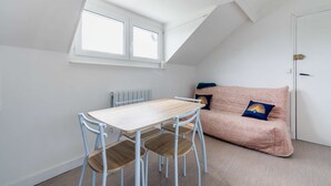 A small, well-lit room with a sloped ceiling, a table with chairs, a radiator, and a sofa with patterned cushions. - Apartment 300m from the beach - Shared outdoor area (Asnelles)