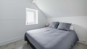 A bedroom with a sloped ceiling, a single bed with grey bedding, a bedside table with a lamp, and a window with a view. - Apartment 300m from the beach - Shared outdoor area (Asnelles)