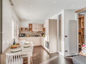 Une cuisine moderne avec des meubles blancs, une table à manger pour quatre personnes et une porte-fenêtre coulissante donnant sur un espace de rangement.