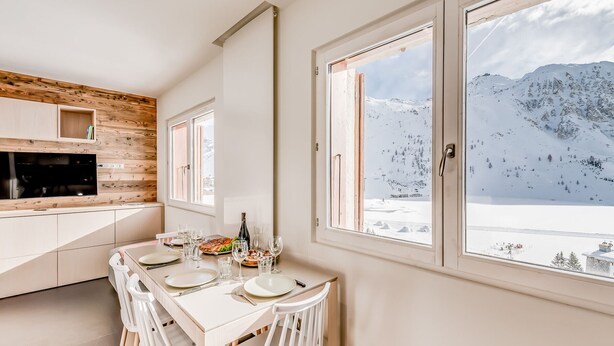 A modern kitchen with wooden accents, a dining area with white furniture, and large windows offering a mountain view. - Renovated apartment for 4, lake view, hotel services (Tignes)