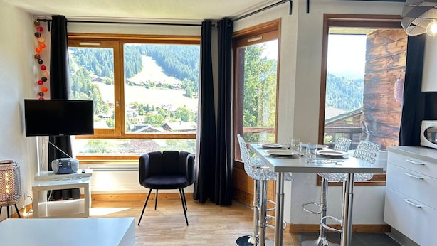 A modern kitchen with a dining area, large windows with a scenic view, a flat-screen TV, and a bar stool. - Renovated Apartment, Walking Distance to Slopes, South-West Balcony, Comfort Amenities (Les Gets)
