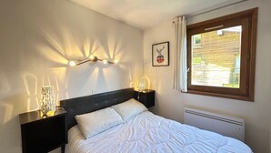A bedroom with a bed, bedside tables, a wall-mounted light fixture, a window with curtains, and a framed picture. - Renovated Apartment, Walking Distance to Slopes, South-West Balcony, Comfort Amenities (Les Gets)