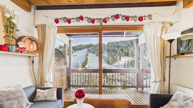 Ein gemütliches Zimmer mit Holzbalkendecke, einem Balkon mit Blick auf eine verschneite Landschaft, einem kleinen Tisch mit einer Dekoration und zwei Sofas.