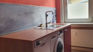 Ett kompakt kök med tvättmaskin, diskho och ett fönster med utsikt över omgivningen.