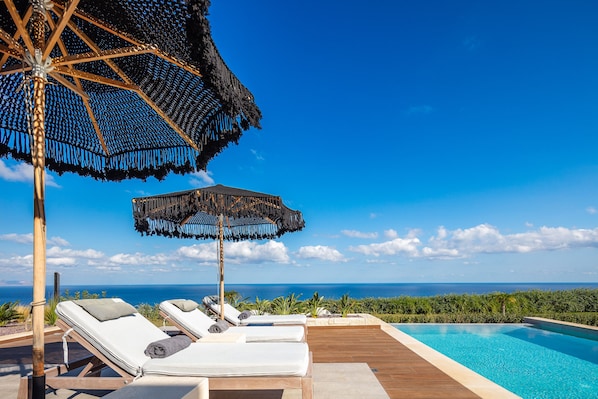 Un espace aménagé au bord de la piscine, avec des chaises longues et un grand parasol, offrant une vue imprenable sur l’océan bleu azur.