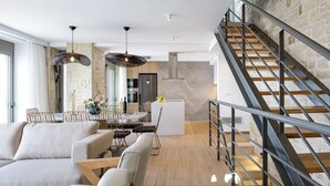 A modern living room with a sofa, dining table, and chairs, a staircase with metal railings, and a kitchen area with a stone wall. - Perfect Hideaway for Families & Unmatched Sunsets (Rethymno)