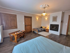 A bedroom with a bed, a wooden bench, a desk, a door, a fireplace, and a ceiling light fixture.