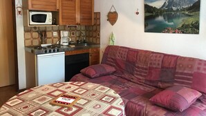A compact living space with a kitchenette, a bed with a patterned blanket, and a painting of a lake and mountains on the wall. - Studio for 2 close to Samoëns center (Samoëns)