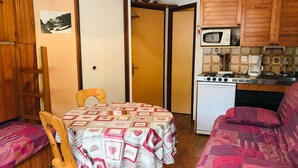 A compact kitchen with a stove, microwave, and cabinets. There is a dining table with a patterned tablecloth and a wooden chair. A sofa is visible in the background. - Studio for 2 close to Samoëns center (Samoëns)