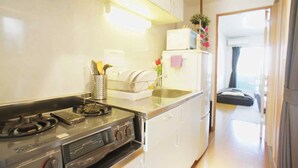Une cuisine compacte équipée d’une cuisinière, d’un évier et d’un égouttoir. On aperçoit un réfrigérateur à l’arrière-plan, et on distingue un lit à travers une porte ouverte.