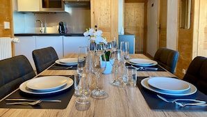A dining area with a wooden table set for four, surrounded by chairs. A vase with white flowers is placed in the center.