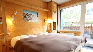 A wooden room with a bed, a window with a view of greenery, and a wall-mounted picture of a ski lift.
