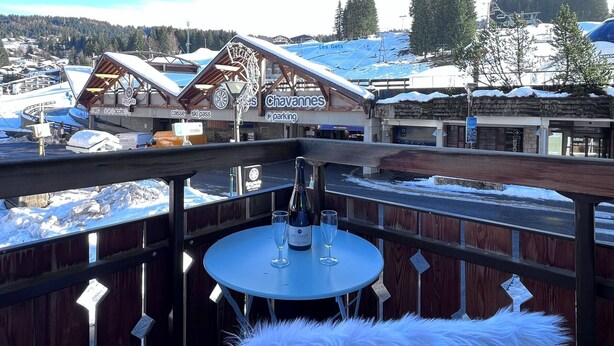 A balcony with a table and chairs, overlooking a snowy village with buildings and a ski lift.