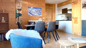 A cozy room with a wooden wall, a dining table with chairs, a small kitchen area, and a bookshelf.
