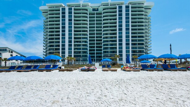 A high-rise hotel with multiple balconies, a beach area with sun loungers and umbrellas, and a clear blue sky.